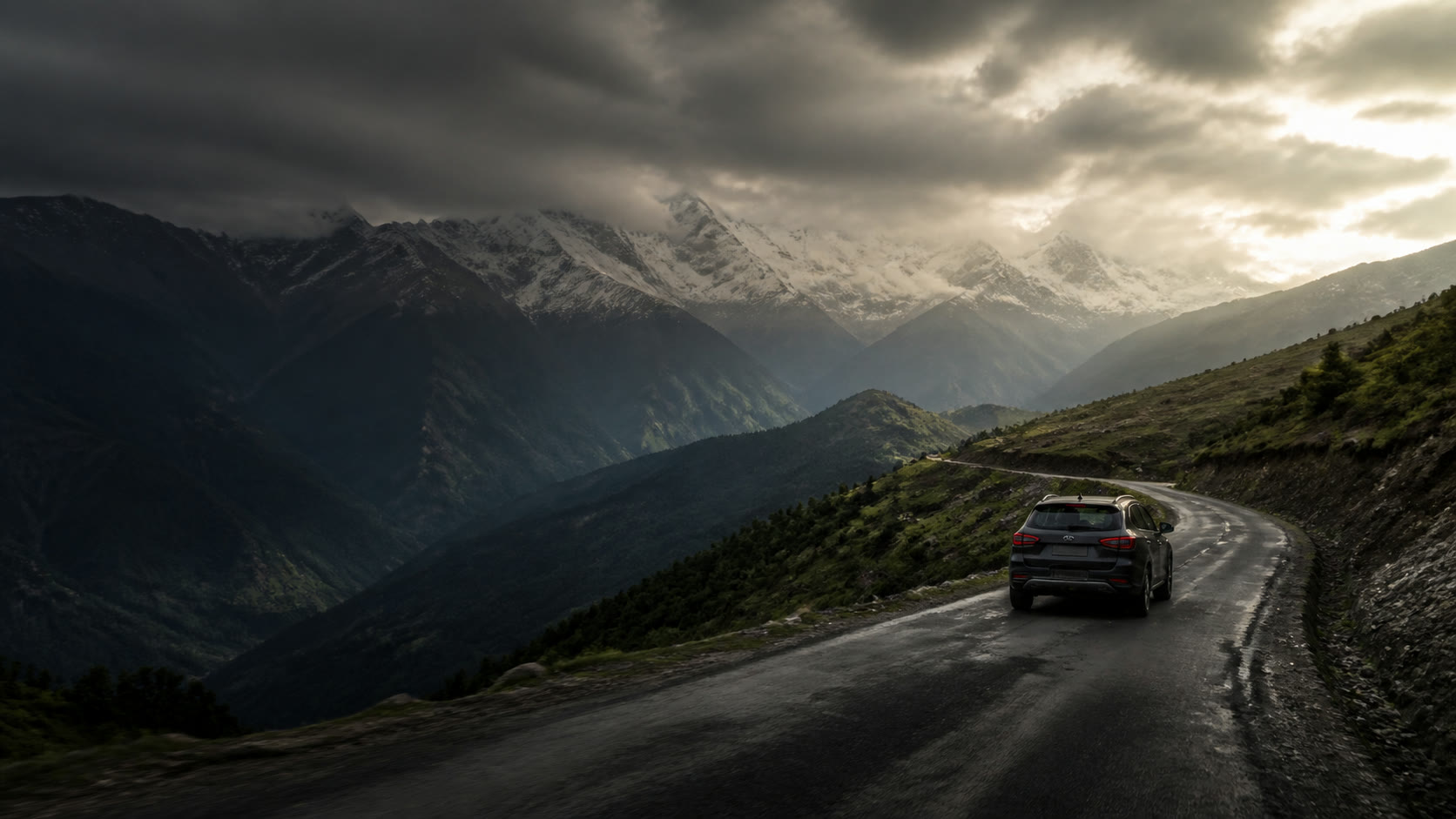 A graphite SUV following a mountain road beneath the Himalayas